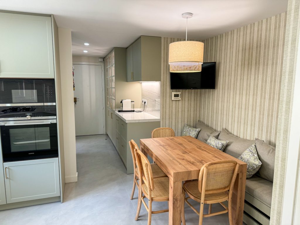 Cocina reformada en Sant Gervasi con muebles verde salvia, zona de comedor con banco y mesa de madera, y lámpara colgante de diseño natural.